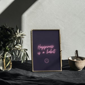 Op de afbeelding: Een gouden ingelijste print met een donkerpaarse achtergrond en roze neontekst die "Happiness is a habit" luidt met een roze smiley onder de tekst.