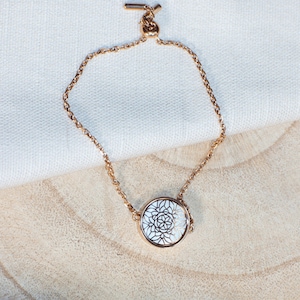 Pode incluir: Um bracelete de corrente dourada com um pingente circular branco com um design de mandala detalhado.