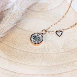 Könnte beinhalten: Eine roségoldene Halskette mit einem runden Anhänger mit einem floralen Design aus Perlmutt. Ein kleiner herzförmiger Anhänger mit Perlmutt-Inlay hängt neben dem Anhänger. Die Halskette wird auf einer hellbraunen Holzoberfläche präsentiert.