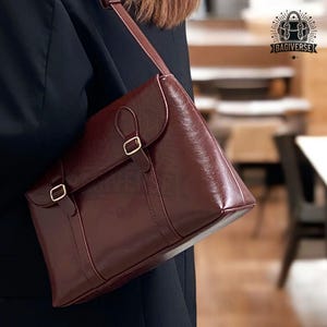 May include: A burgundy leather satchel bag with a long strap and gold buckles. The bag has a flap closure and a structured shape.