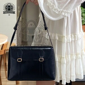 May include: A black leather satchel bag with a long shoulder strap and two gold buckles. The bag has a flap closure and a structured shape.