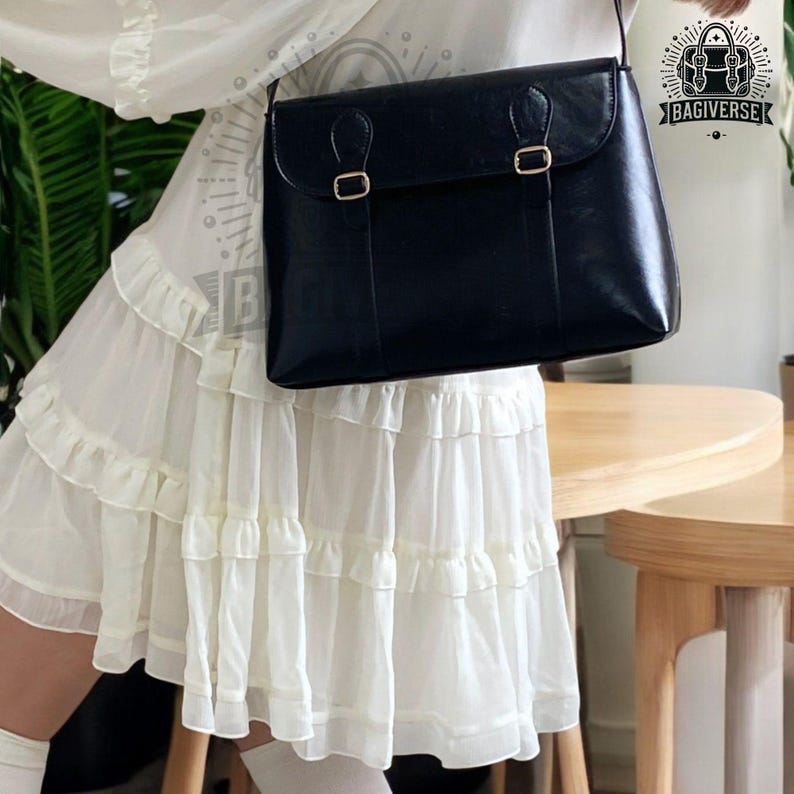 May include: A black leather satchel bag with gold buckles and a strap. The bag is worn by a person wearing a white ruffled skirt.