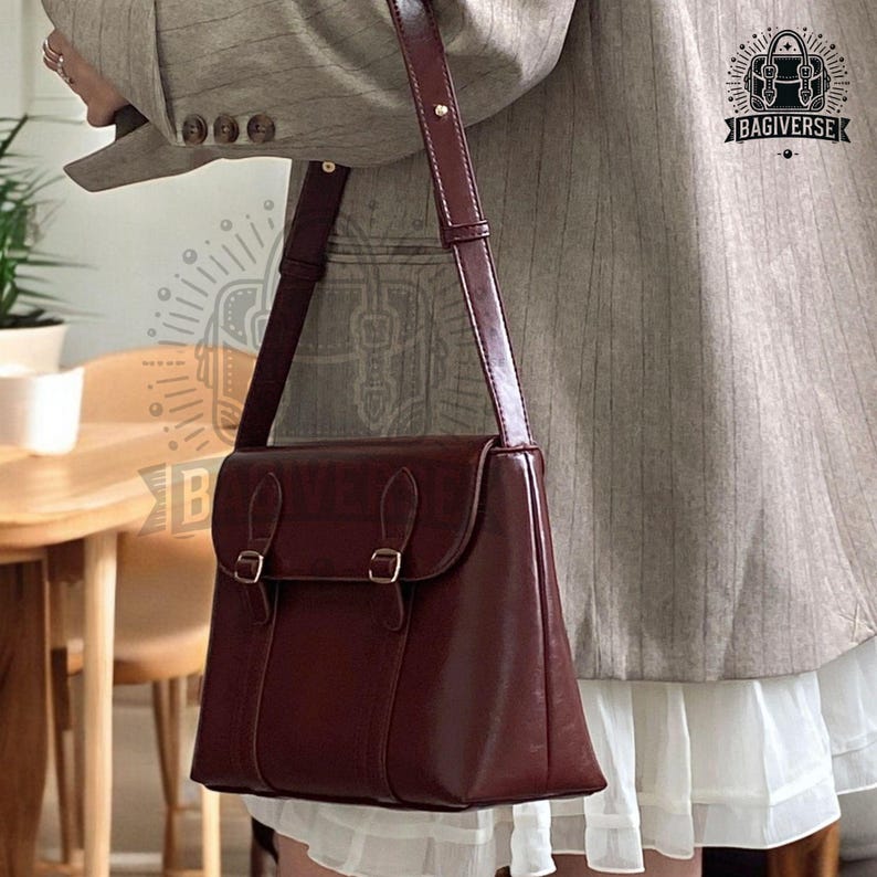 May include: A burgundy leather satchel bag with a long adjustable strap. The bag has two buckles on the front flap and a single compartment.
