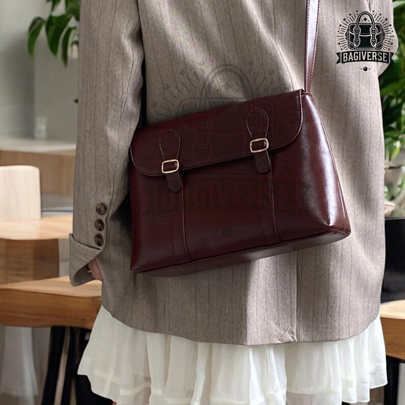 May include: A brown leather satchel bag with a flap closure and two gold buckles. The bag has a long adjustable strap and is worn over the shoulder.
