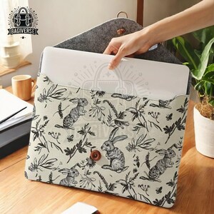 Puede incluir: Una funda para portátil blanca con un patrón de conejo y flores en blanco y negro. La funda está hecha de un tejido suave y duradero y tiene un cierre de cremallera. La funda se muestra con un portátil en su interior.