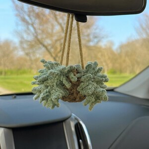 May include: A hanging car air freshener featuring a crocheted plant design. The plant is a light green color, and the pot is a brown color. The air freshener hangs from a natural-colored rope, suspended from the rearview mirror.