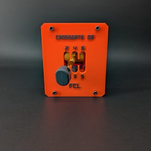 May include: A bright orange rectangular gear shifter gate with a dark gray knob. The plate features the text CROSSGATE 03 at the top, gear numbers 2, 4, 6, 3, 5, and the letters FCL at the bottom. It is secured by four black corner screws.