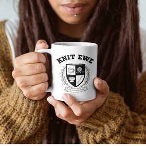 Puede incluir: Taza de cerámica blanca con un diseño en blanco y negro. El diseño presenta un escudo con una taza de café, una bola de hilo, agujas de tejer y un libro. El texto "Knit Ewe" y "Don't Fret, Be Yarn" están en la taza.