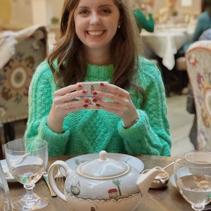 Könnte beinhalten: Eine Frau in einem grünen Pullover hält eine kleine weiße Teetasse mit rotem Nagellack. Eine Teekanne mit blau-weißem Design steht auf einem Holztisch mit einem Glas Wasser und einer silbernen Zuckerdose.