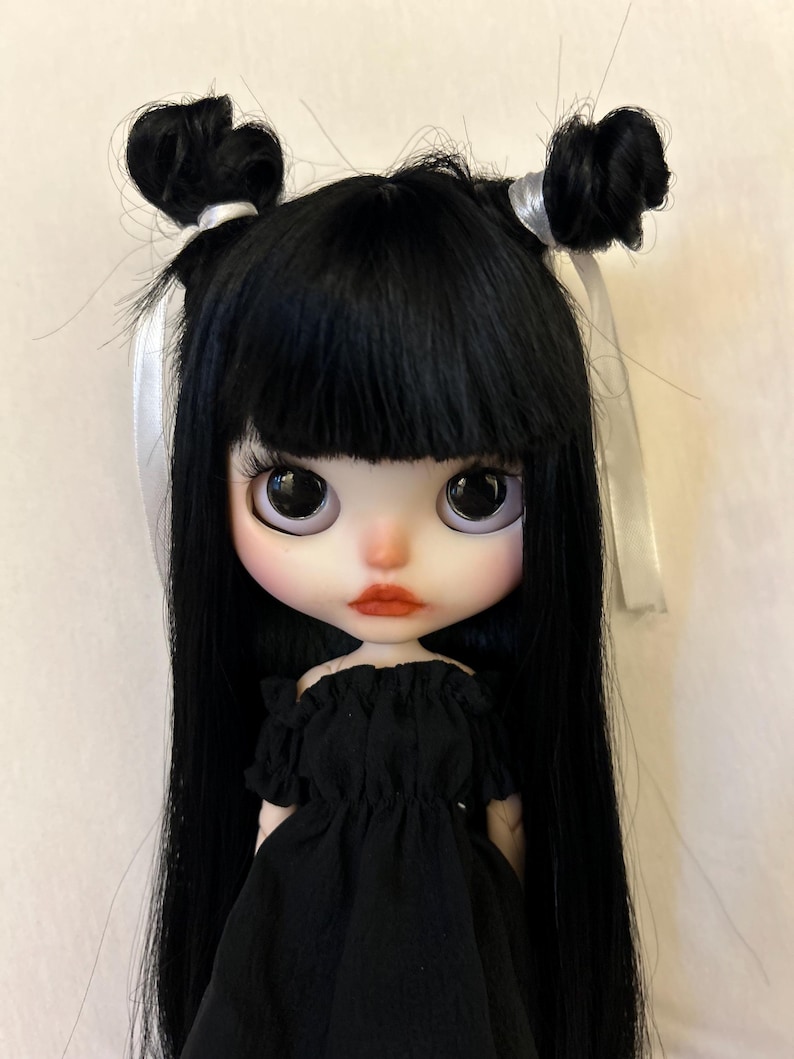 Puede incluir: Una mu&ntilde;eca de pelo negro con cintas blancas en el pelo, que lleva un vestido negro de hombros descubiertos. La mu&ntilde;eca tiene ojos grandes y oscuros y labios rojos.