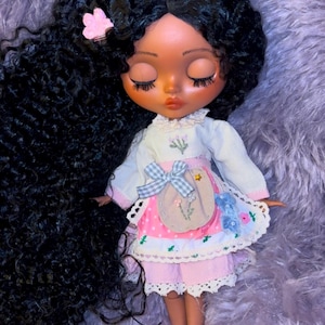Puede incluir: Una mu&ntilde;eca con el pelo largo y negro rizado, que lleva un vestido rosa y blanco con estampado floral. La mu&ntilde;eca tiene la piel morena y los ojos cerrados.