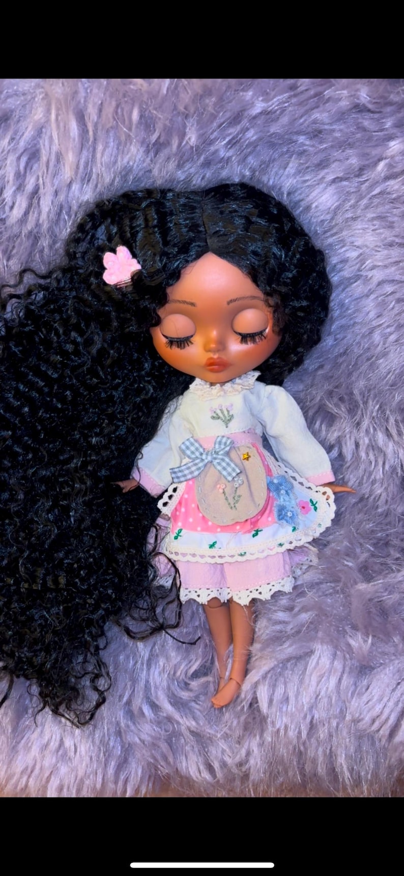 Puede incluir: Una mu&ntilde;eca con el pelo largo y negro rizado, que lleva un vestido rosa y blanco con estampado floral. La mu&ntilde;eca tiene la piel morena y los ojos cerrados.