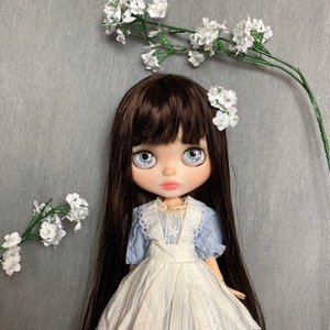 Puede incluir: Una mu&ntilde;eca con el pelo largo y casta&ntilde;o, ojos azules y un vestido blanco y azul. La mu&ntilde;eca lleva un delantal blanco y tiene flores blancas en el pelo. La mu&ntilde;eca est&aacute; de pie sobre un fondo gris.