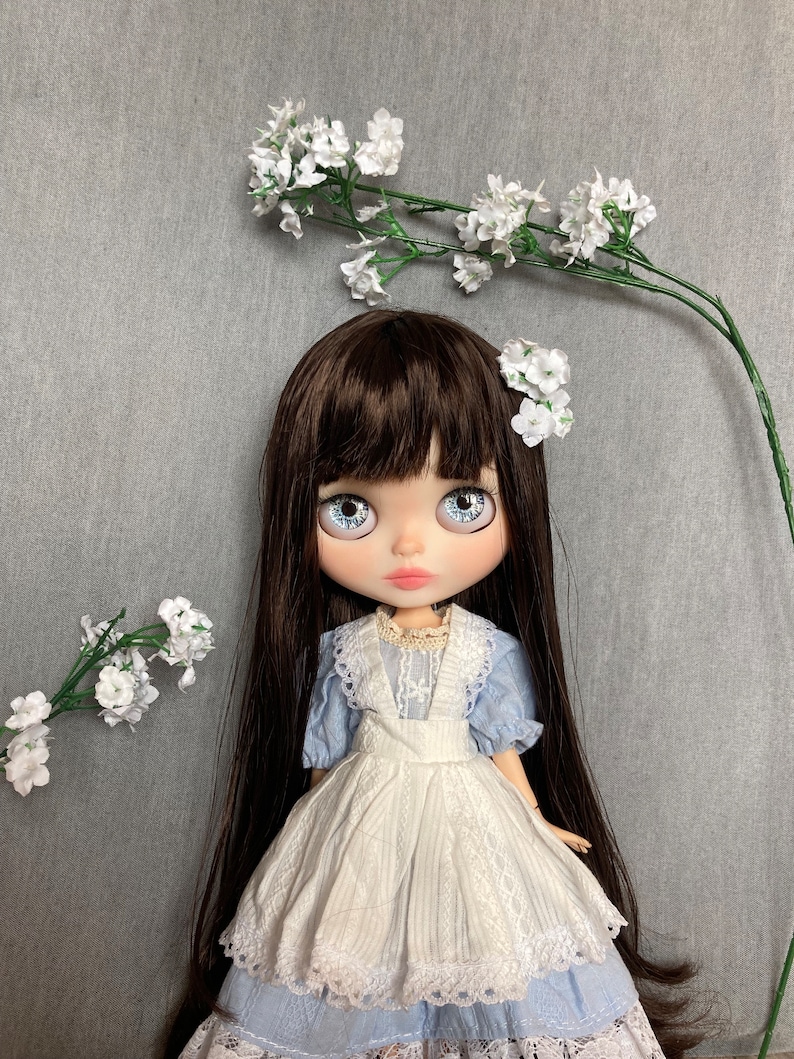 Puede incluir: Una mu&ntilde;eca con el pelo largo y casta&ntilde;o, ojos azules y un vestido blanco y azul. La mu&ntilde;eca lleva un delantal blanco y tiene flores blancas en el pelo. La mu&ntilde;eca est&aacute; de pie sobre un fondo gris.