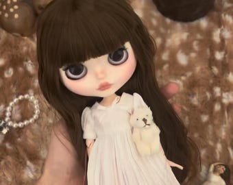 Può includere: Una bambola con lunghi capelli castani e frangia, che indossa un vestito bianco a maniche corte con gonna plissettata. La bambola ha grandi occhi scuri e tiene in mano un piccolo orsacchiotto bianco. Indossa collant a righe rosa chiaro e ha la carnagione chiara.