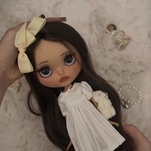 Può includere: Una bambola con lunghi capelli castano scuro, grandi occhi grigi e carnagione chiara. La bambola indossa un vestito bianco plissettato a maniche corte e un fiocco giallo pallido tra i capelli. Tiene in mano un piccolo peluche bianco.