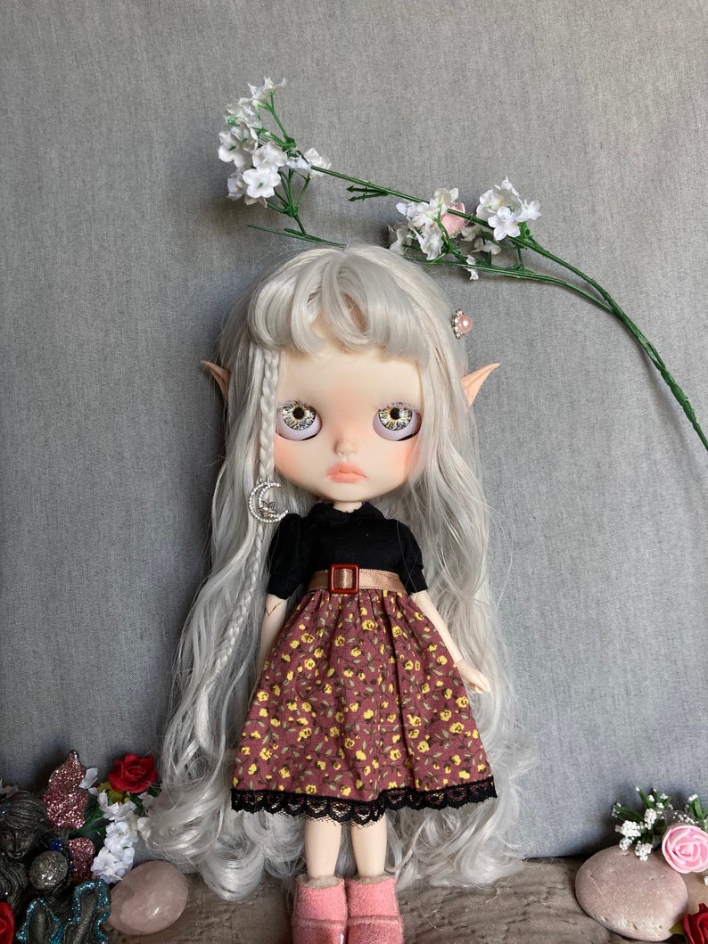 Puede incluir: Una mu&ntilde;eca con el pelo largo y blanco que lleva un vestido negro y marr&oacute;n con estampado floral. La mu&ntilde;eca tiene ojos grandes y azules y lleva un cintur&oacute;n marr&oacute;n con una hebilla plateada.