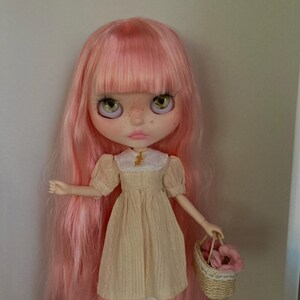Puede incluir: Una mu&ntilde;eca con el pelo rosa que lleva un vestido de color crema con un cuello blanco y una peque&ntilde;a cesta de flores rosas.