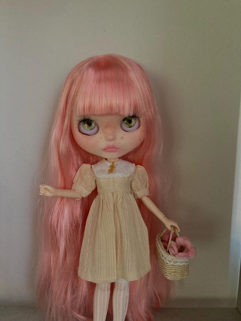 Puede incluir: Una mu&ntilde;eca con el pelo rosa que lleva un vestido de color crema con un cuello blanco y una peque&ntilde;a cesta de flores rosas.