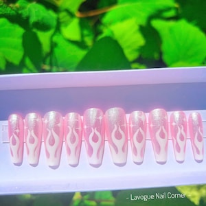 May include: Pink and white press-on nails with a flame design and glitter accents. The nails are displayed in a white box with the text "Lavogue Nail Corner".