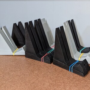 以下が含まれることがあります： 黒とライトグレーのプラスチック製L字型スタンドのコレクション。それぞれ垂直のバックサポートと水平のベースを備えています。複数のスタンドがグループ化されており、各ユニットのベースにはカラフルな輪ゴムが巻き付けられています。