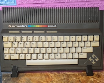 Può includere: Un computer domestico Commodore Plus/4 grigio scuro con tastiera QWERTY beige, tasti funzione, una striscia con logo arcobaleno e un gruppo di tasti freccia sul lato destro. Il dispositivo è sostenuto da due supporti in plastica nera.