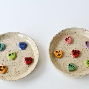 May include: Two speckled ceramic plates each hold several heart-shaped candies. The candies are various colors, including blue, green, orange, and red, with phrases like "AS IF!" and "HARD PASS" written on them. The plates are labeled A and B.
