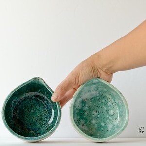 May include: Two teardrop-shaped ceramic bowls with a glossy finish. Bowl B features a deep emerald green glaze with dark speckles, while bowl C displays a lighter mint green glaze with white mottled patterns and dark flecks throughout the surface.