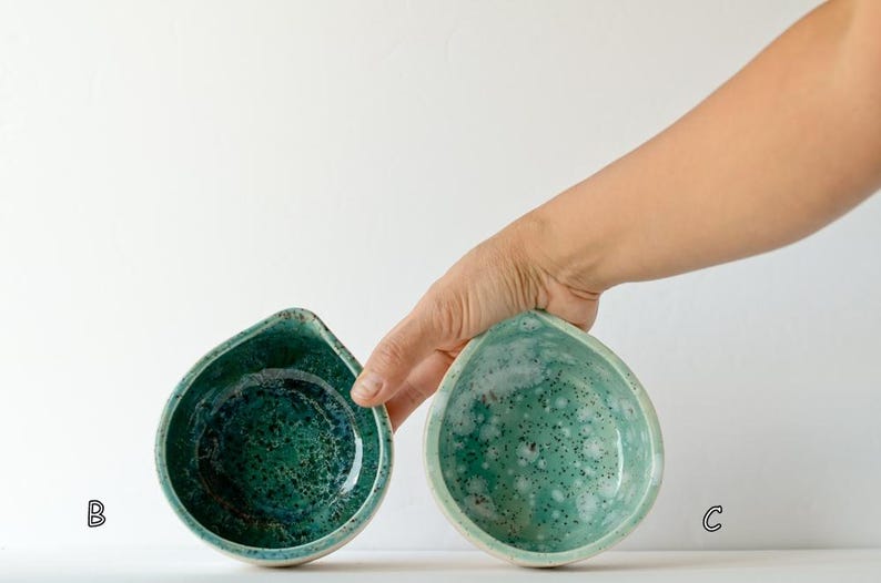 May include: Two teardrop-shaped ceramic bowls with a glossy finish. Bowl B features a deep emerald green glaze with dark speckles, while bowl C displays a lighter mint green glaze with white mottled patterns and dark flecks throughout the surface.