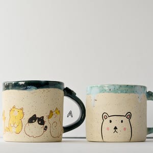 May include: Two ceramic mugs with unique designs. One mug features a group of cartoon cats in yellow, black, and white. The other mug has a bear illustration. Both mugs have speckled beige bodies and dark green or blue rims and handles.