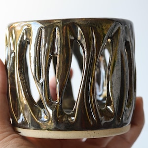 Puede incluir: Un macetero de cerámica marrón con un diseño único, ondulado y recortado. El macetero tiene un esmalte blanco en el borde inferior.