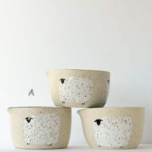 May include: Three ceramic bowls with a speckled cream finish, each featuring a hand-painted white sheep design with black details. The bowls are arranged in a stack, with one bowl labeled A on top, and two bowls labeled B and C at the base.