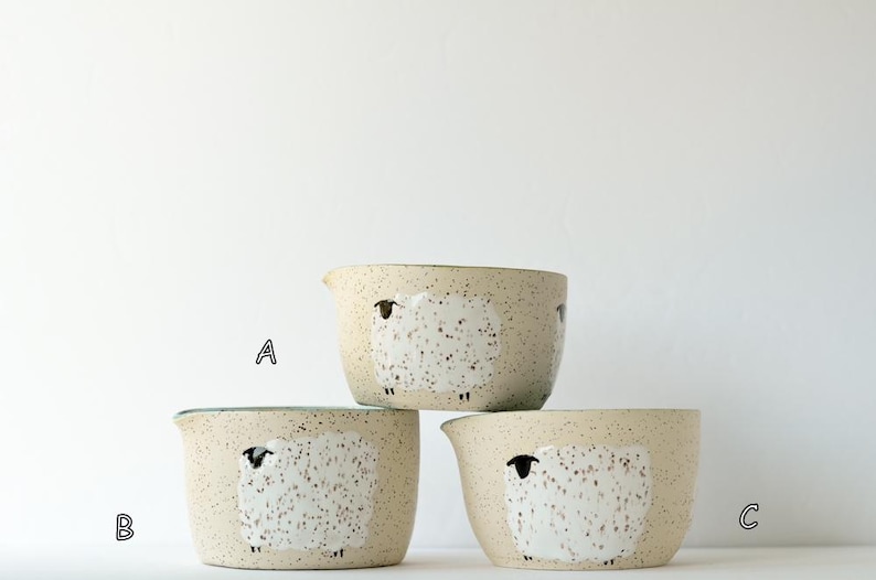 May include: Three ceramic bowls with a speckled cream finish, each featuring a hand-painted white sheep design with black details. The bowls are arranged in a stack, with one bowl labeled A on top, and two bowls labeled B and C at the base.