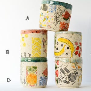 May include: Five ceramic cups stacked in a pyramid, each featuring a speckled cream base and unique colorful illustrations. Cup A shows ice cream and plants, B displays lemons and flamingos, C features bananas and berries, D shows citrus slices, and E depicts sewing supplies.