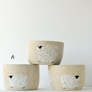 May include: Three ceramic bowls with a speckled cream finish, each featuring a hand-painted white sheep illustration. The bowls are arranged in a stack, with one bowl on top and two below, labeled with the letters A, B, and C in black font.