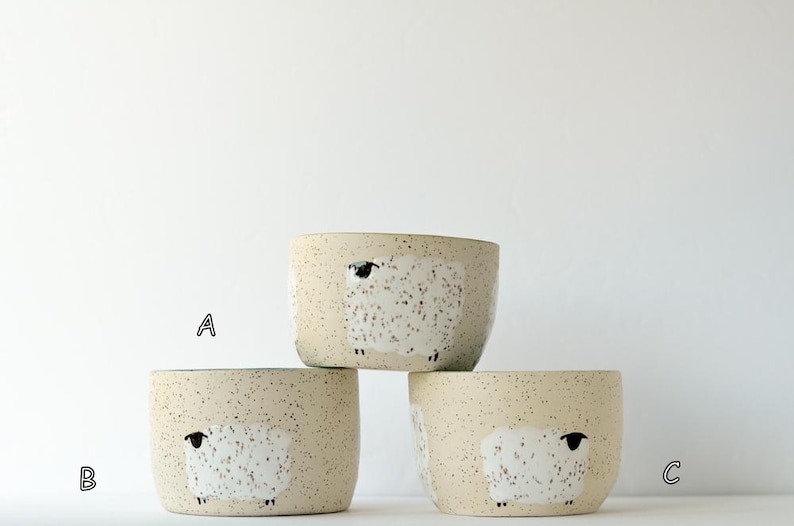 May include: Three ceramic bowls with a speckled cream finish, each featuring a hand-painted white sheep illustration. The bowls are arranged in a stack, with one bowl on top and two below, labeled with the letters A, B, and C in black font.