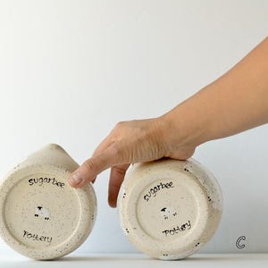 May include: Two speckled cream-colored ceramic pottery pieces, each featuring a hand-stamped sheep icon and the text "sugarbee pottery" on the base. The items are labeled with the letters B and C, showcasing the underside of the ceramic vessels.