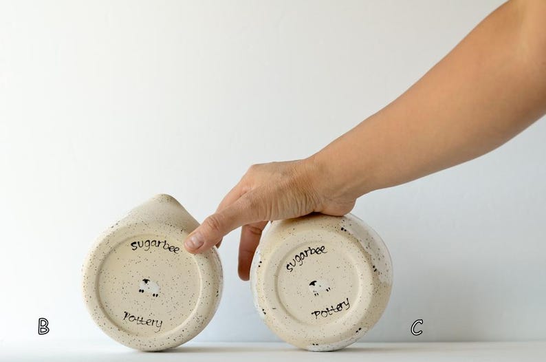 May include: Two speckled cream-colored ceramic pottery pieces, each featuring a hand-stamped sheep icon and the text "sugarbee pottery" on the base. The items are labeled with the letters B and C, showcasing the underside of the ceramic vessels.