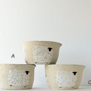May include: Three ceramic bowls with a speckled beige finish, each featuring a white sheep illustration with a black head and legs. The bowls are arranged in a stack, labeled with the letters A, B, and C, and each has a small pouring spout.