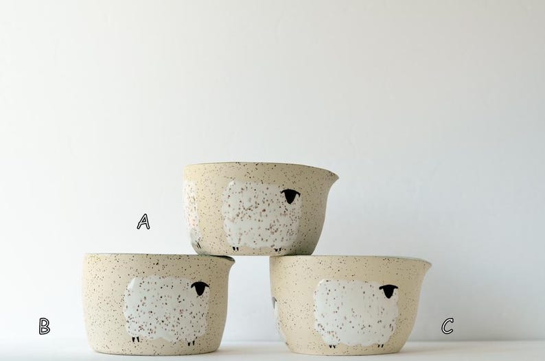 May include: Three ceramic bowls with a speckled beige finish, each featuring a white sheep illustration with a black head and legs. The bowls are arranged in a stack, labeled with the letters A, B, and C, and each has a small pouring spout.