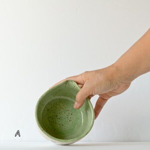 May include: A small, round ceramic bowl with a light green interior featuring dark green speckles and a distinct pouring spout on one side. The exterior of the bowl is a neutral off-white color. The letter A is printed in black near the base.