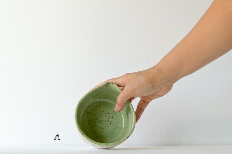 May include: A small, round ceramic bowl with a light green interior featuring dark green speckles and a distinct pouring spout on one side. The exterior of the bowl is a neutral off-white color. The letter A is printed in black near the base.