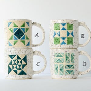 May include: Four ceramic mugs with speckled cream glaze and geometric patterns in shades of blue and green. Each mug features a unique quilt-inspired design labeled with the letters A, B, C, and D in a simple black font next to each handle.