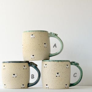 Puede incluir: Tres tazas de cerámica con un acabado crema moteado y un patrón repetitivo de pequeñas ovejas blancas y negras. Cada taza presenta un asa y un borde de color verde azulado, con las letras A, B y C visibles junto a cada taza.