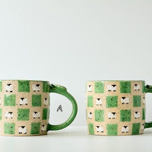 May include: Two ceramic mugs featuring a green and beige checkered pattern with small white and black sheep illustrations on the beige squares. Each mug has a green handle and the letters A and B are printed next to the respective mugs.