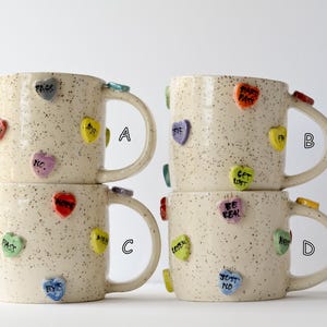 May include: Four speckled ceramic mugs stacked, each adorned with colorful heart-shaped embellishments. The hearts feature phrases like "BYE," "NO," and "GET LOST." The mugs have large handles and are in a neutral color.