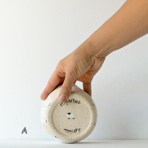 May include: A round, cream-colored ceramic bowl with dark speckles, featuring the text sugarbee Pottery and a small sheep illustration on the bottom. The letter A is printed to the left of the bowl, which has a smooth, rounded base.