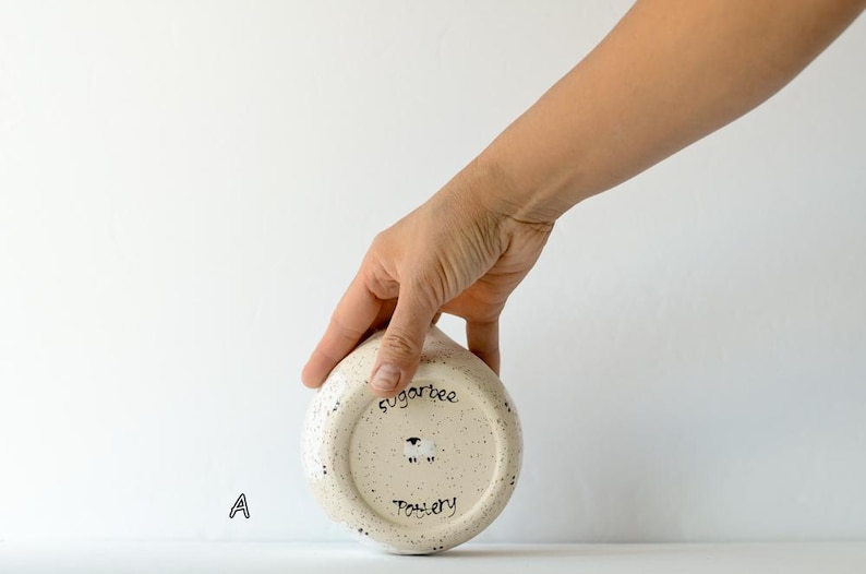 May include: A round, cream-colored ceramic bowl with dark speckles, featuring the text sugarbee Pottery and a small sheep illustration on the bottom. The letter A is printed to the left of the bowl, which has a smooth, rounded base.