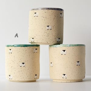 Könnte beinhalten: Drei zylindrische Keramikbecher mit gesprenkelter cremefarbener Oberfläche und einem wiederkehrenden Muster aus kleinen weißen Schafen mit schwarzen Gesichtern. Jeder Becher hat einen farbigen Rand, gekennzeichnet mit A, B und C.