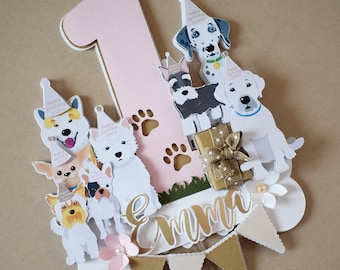 Puede incluir: Un adorno para pastel de color rosa y blanco con un gran número uno, una pequeña caja de regalo y varios perros de dibujos animados con sombreros de fiesta. Los perros son de diferentes razas, incluyendo un dálmata, un schnauzer y un terrier. El adorno también incluye una pancarta con el nombre "Emma" en letras doradas.
